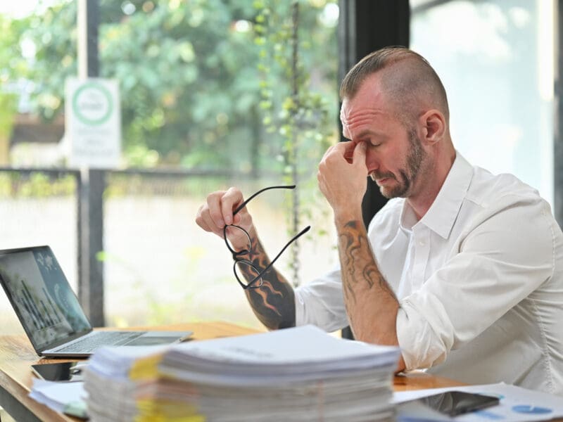 Overworked caucasian man worker holding glasses and rubbing nose suffering from chronic dry eye syndrome bridge.