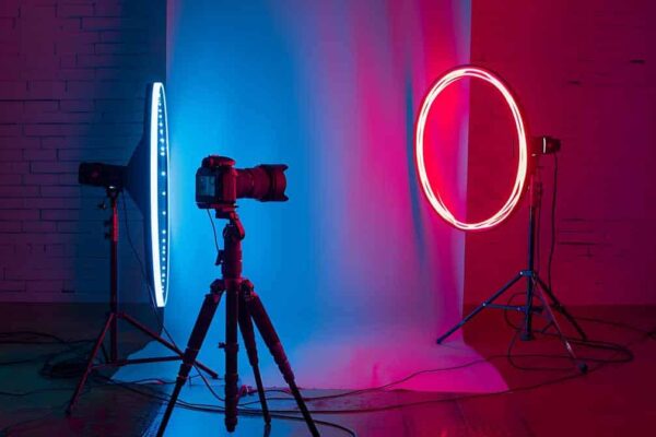 A ring light with an LED neon lamp on a tripod, flanked by a DSLR camera on another tripod, in a studio with moody blue and red lighting